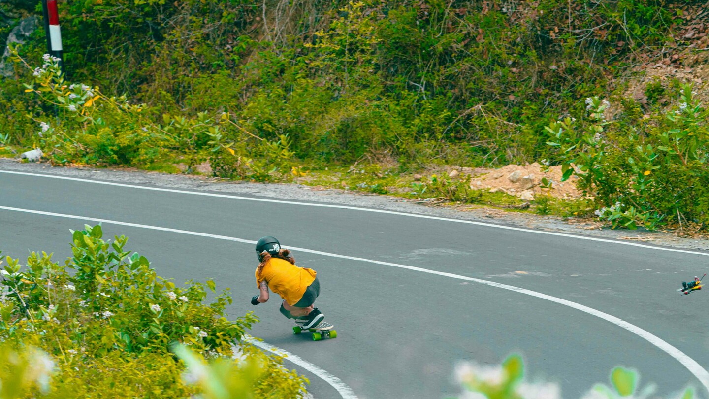 Anna Pixner, a downhill skateboarder, is riding in a crouched position through a tight curve on a rural road. She is wearing a black helmet, yellow T-shirt, and protective gear. The surroundings are filled with dense vegetation.