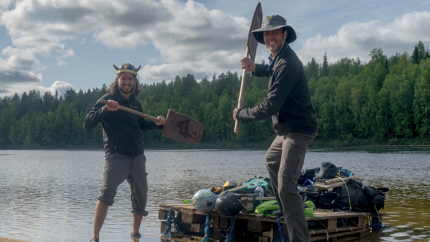 Two men are playfully posing with improvised paddles on the shore of a lake. The man on the left is wearing a Viking helmet, while the man on the right has a wide-brimmed hat. They stand next to a makeshift raft made of wooden pallets, loaded with gear. In the background, a forested shoreline is visible.